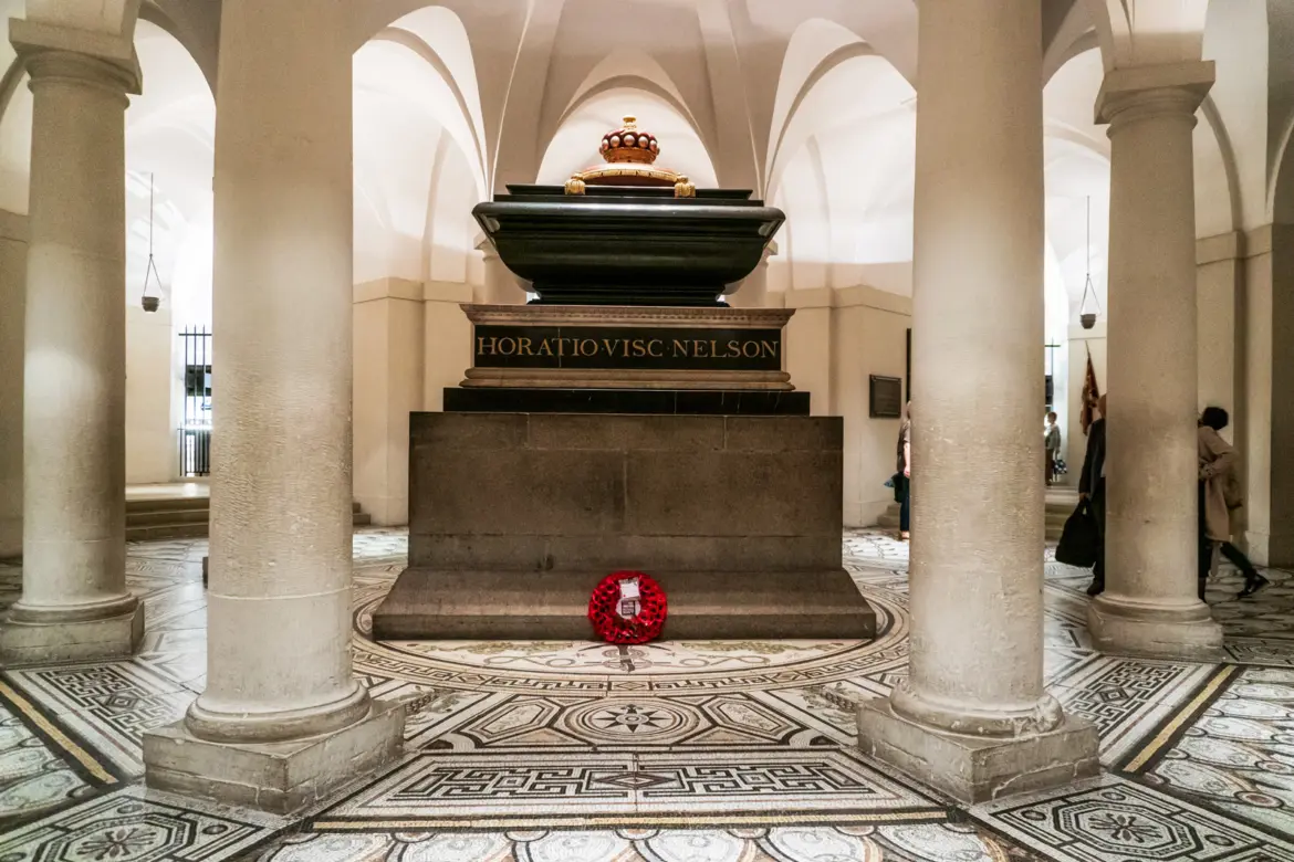 The tomb of Vice-Admiral Lord Nelson at the centre of the crypt at St Paul's Cathedral, with the polished black marble sarcophagus on a granite base, a red poppy wreath at its foot, and the geometric mosaic floor of the Nelson Chamber radiating outward beneath the cathedral's central dome above