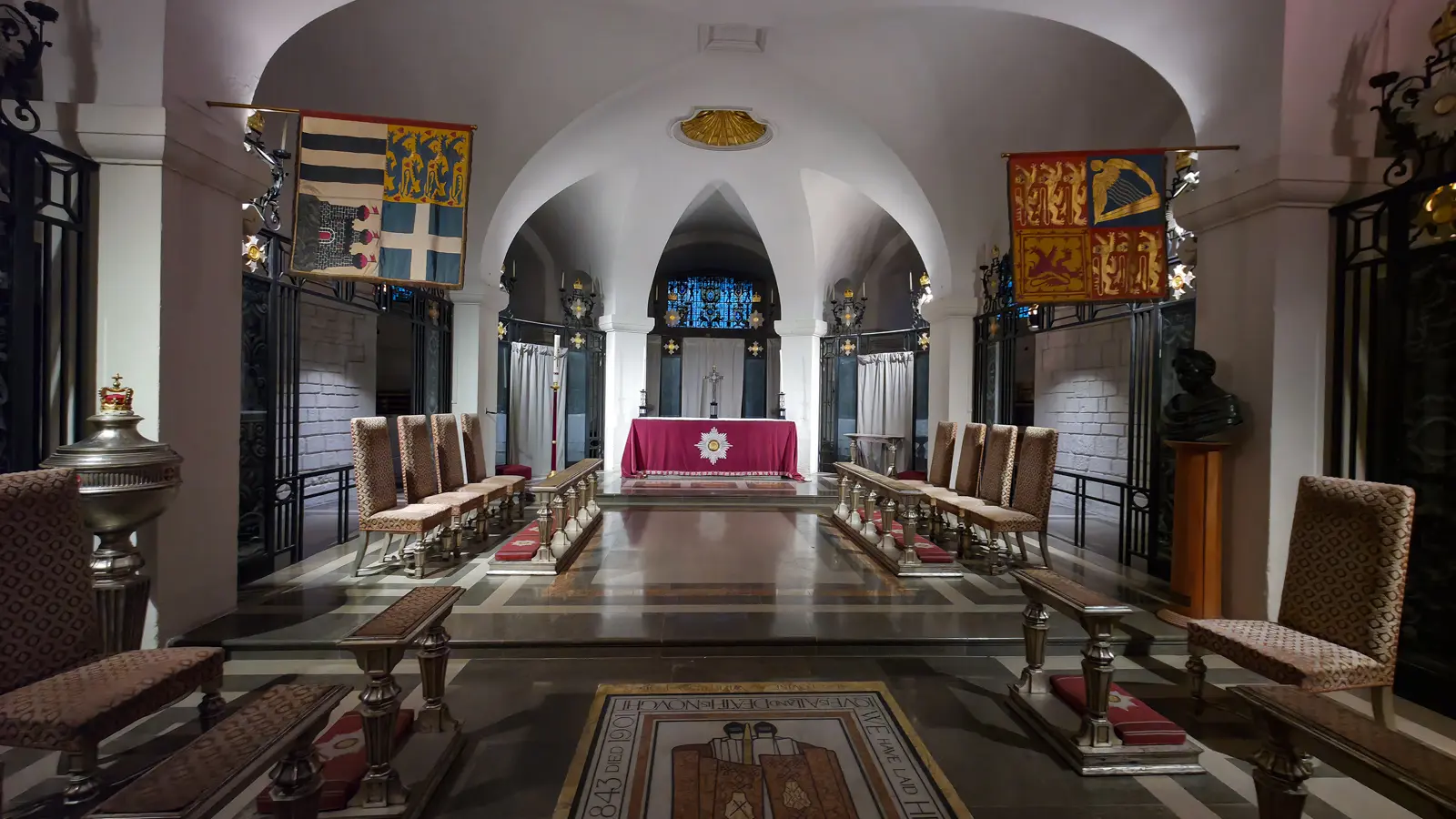 The Order of the British Empire Chapel at the east end of the crypt at St Paul's Cathedral, looking toward the altar with its red cloth and gold OBE star, with heraldic banners of senior members hanging to either side, two rows of upholstered chairs facing in, and a stained-glass window behind the altar