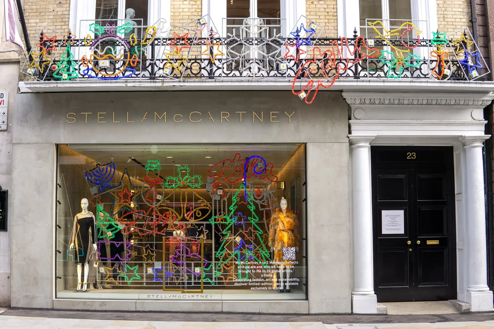The Stella McCartney flagship storefront at 23 Old Bond Street in Mayfair, London with a colourful holiday window display featuring neon-style lit Christmas trees, stars, and seasonal motifs, plus mannequins in the window