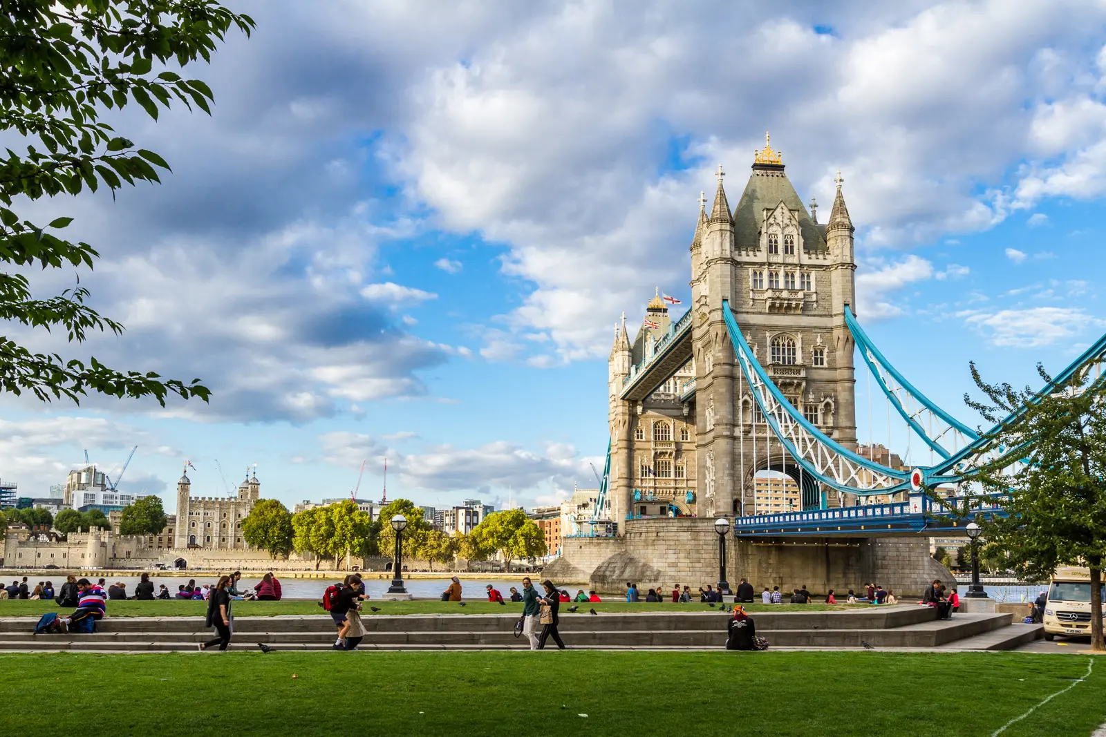Pedestrian promenade and riverside green spaces near Tower Bridge along the River Thames in London, showcasing the vibrant waterfront atmosphere surrounding one of London's most iconic landmarks