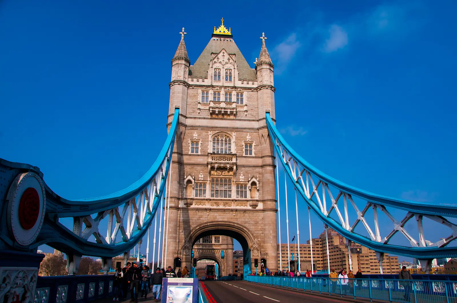 Street-level view while crossing Tower Bridge in London, showcasing the bridge's soaring Gothic Revival towers and distinctive blue suspension elements above the River Thames
