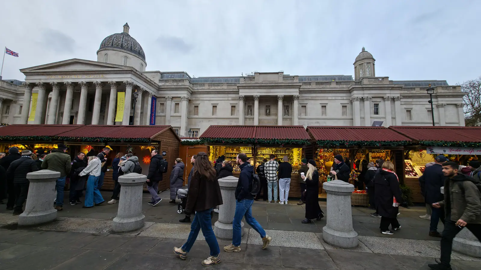 The Trafalgar Square Christmas Market on the National Gallery terrace in December, with around forty wooden alpine-style chalets with red awnings and evergreen garlands, the neoclassical gallery façade and dome rising behind, yellow banners hanging from the columns, and crowds of visitors walking past in winter coats