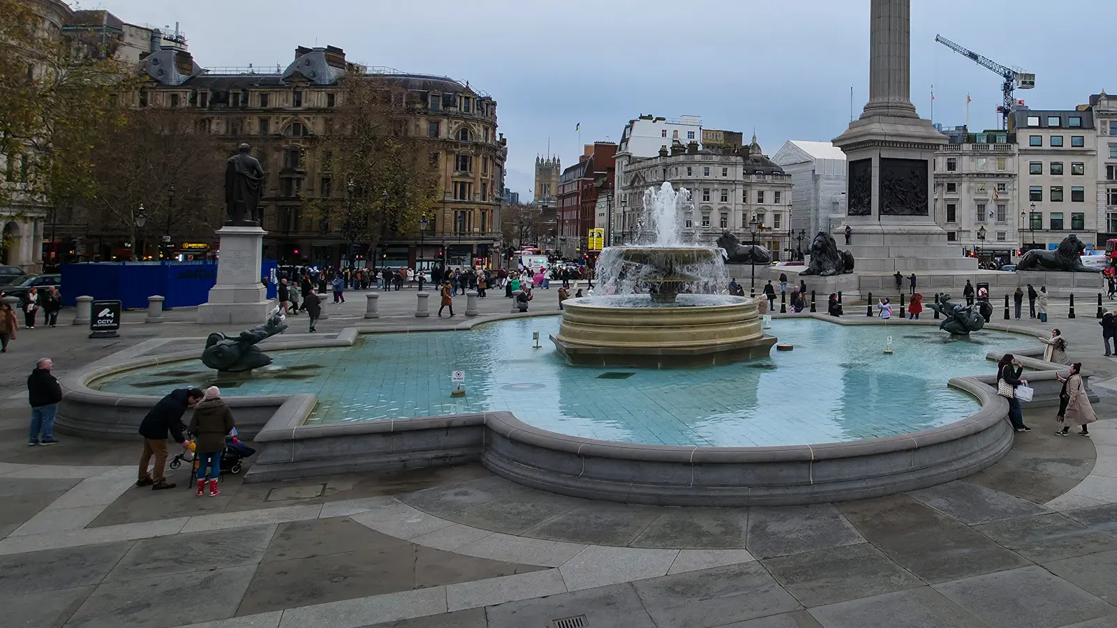 The east fountain of Trafalgar Square on an overcast December afternoon, with the pale blue tile of the basin and the vase-shaped Lutyens central fountain in the foreground, Nelson's Column rising behind, and the Norwegian Christmas tree visible to the right