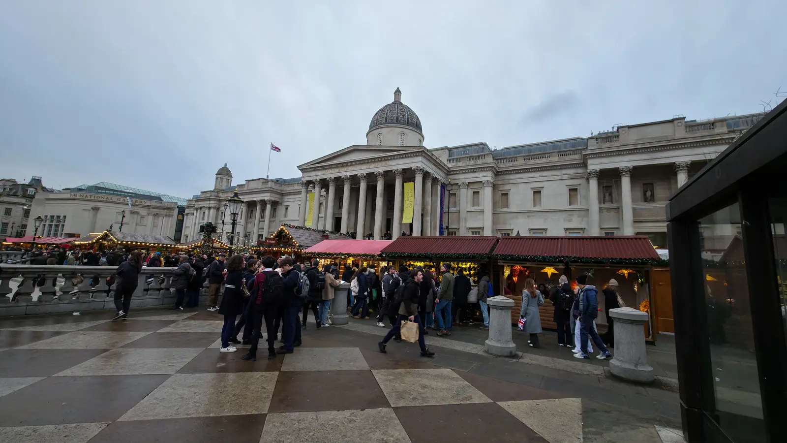 The National Gallery on the north side of Trafalgar Square in December, with its neoclassical façade, central dome, and Corinthian portico, vertical yellow exhibition banners hanging between the columns, and the red-roofed Christmas market chalets running along the upper terrace in the foreground