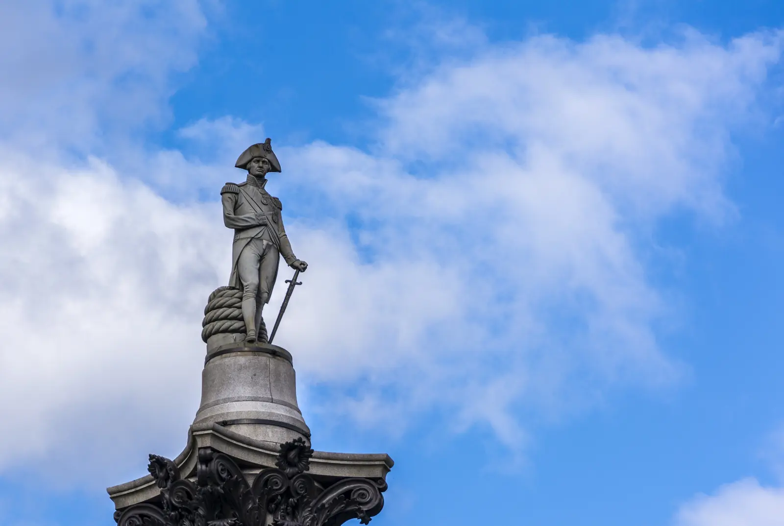 The sandstone statue of Vice-Admiral Lord Nelson at the top of Nelson's Column in Trafalgar Square, in full Royal Navy admiral's uniform with bicorn hat, his right sleeve pinned up after losing the arm at Tenerife in 1797 and a sword held in his left hand, against a blue sky with cumulus clouds