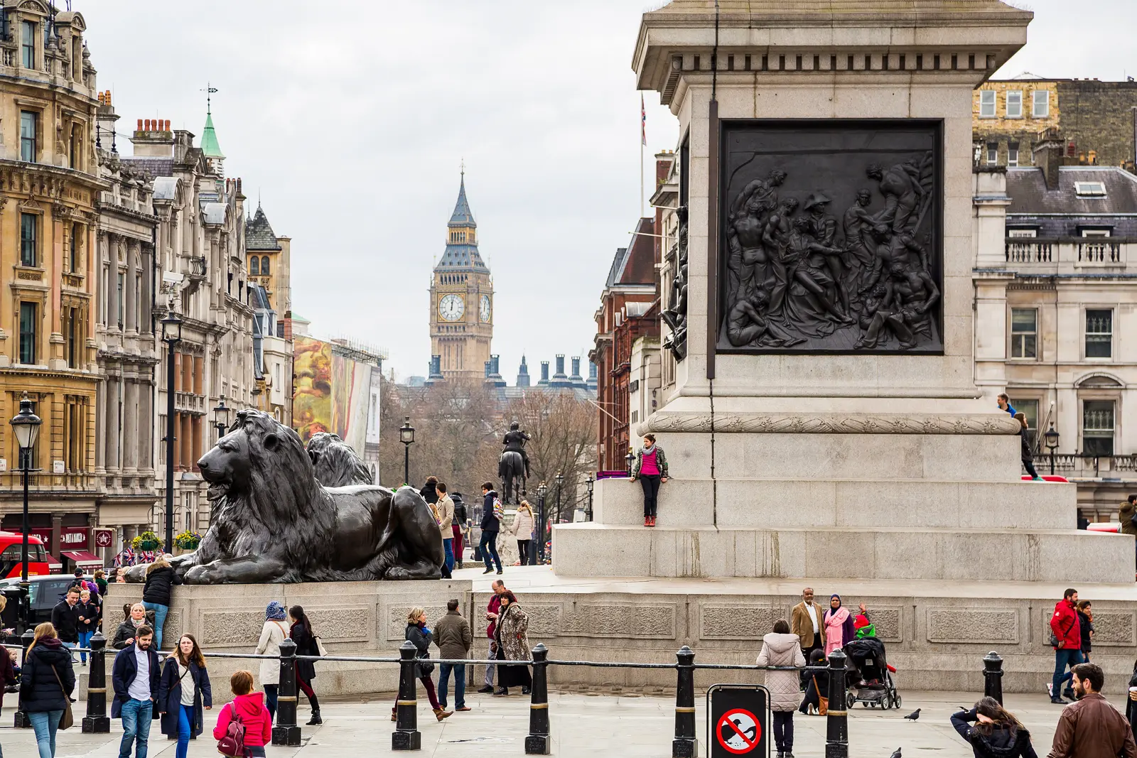 The south side of Nelson's Column at Trafalgar Square showing the bronze Death of Nelson panel mounted on the pedestal, one of the Landseer bronze lions reclining in the left foreground, and the view south down Whitehall toward the clock tower of Big Ben, framed by Victorian London buildings