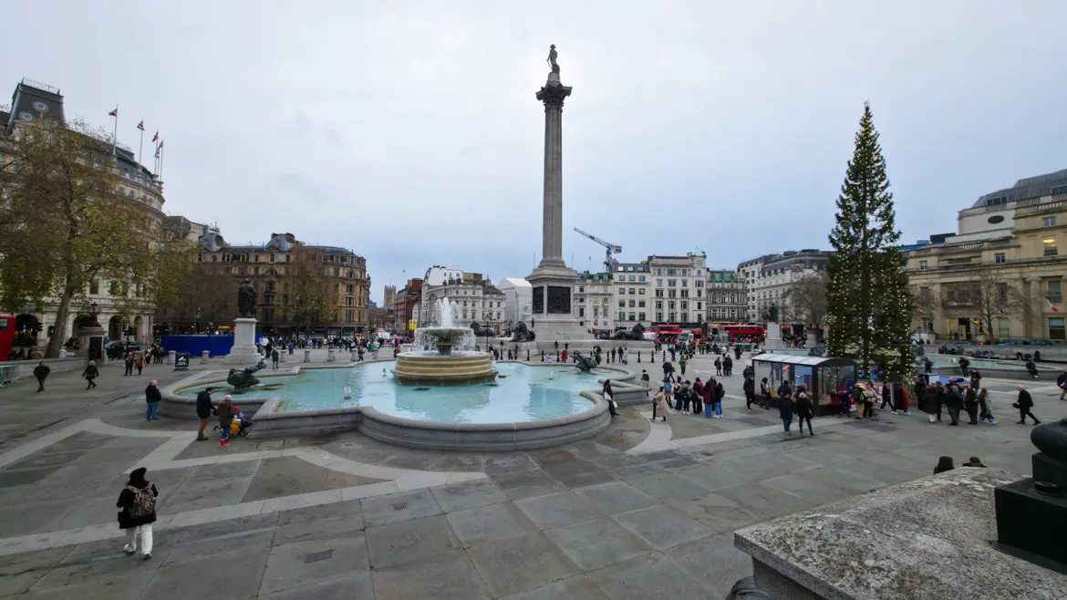 A wide view of Trafalgar Square on a December afternoon from the upper terrace, with Nelson's Column rising in the centre, the east Lutyens fountain in the foreground, the Norwegian Christmas tree lit on the right, and the National Gallery's Sainsbury Wing visible at the right edge under a soft overcast sky