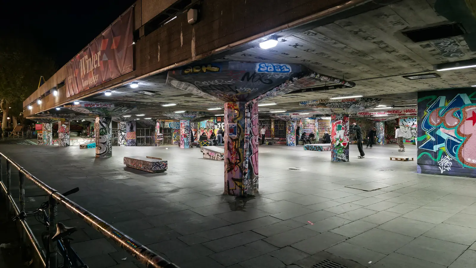 The Undercroft Skate Space beneath the Queen Elizabeth Hall at night, the graffiti-painted concrete columns and ledges lit from above. Skateboarders have used the space since the early 1970s, and the Southbank Centre describes it as the world's longest continually used skate spot. It was saved from redevelopment in 2014 after the Long Live Southbank campaign, with a major restoration completed in 2019