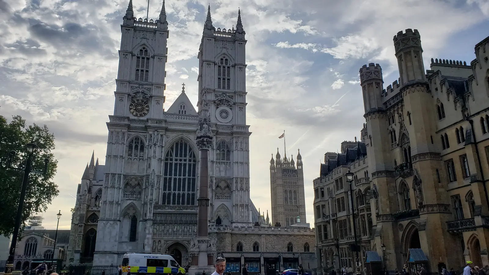 The west front of Westminster Abbey in London with its twin Gothic towers designed by Nicholas Hawksmoor, the central rose window, and Methodist Central Hall and Victoria Tower visible to the right under streaked clouds