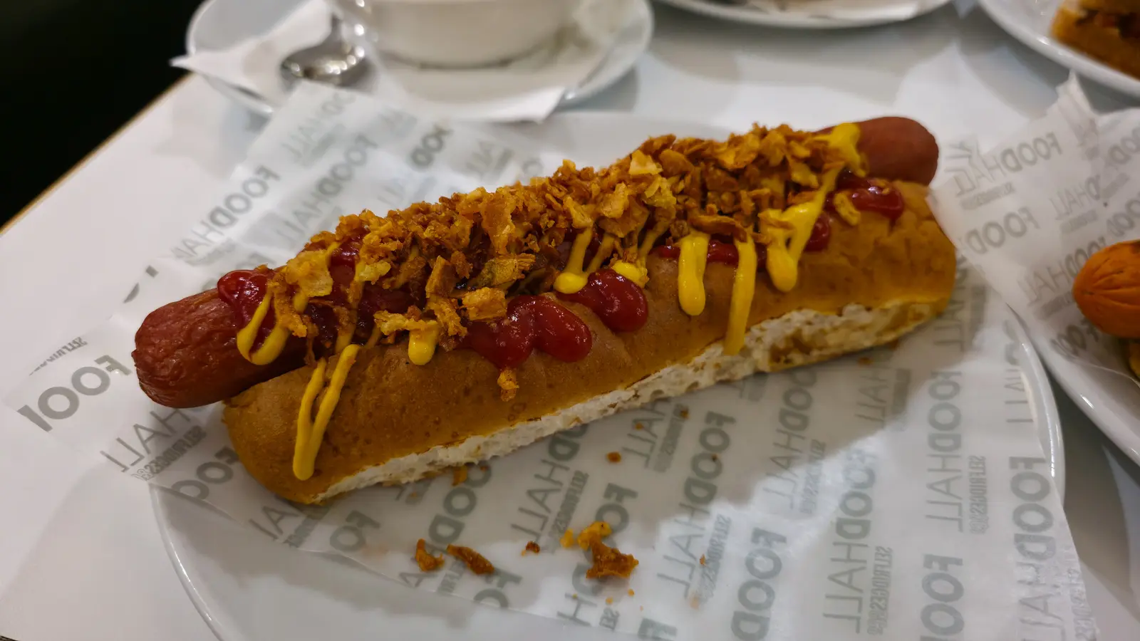 A long beef hot dog in a brioche bun topped with crispy onions, ketchup and mustard on Selfridges Food Hall paper