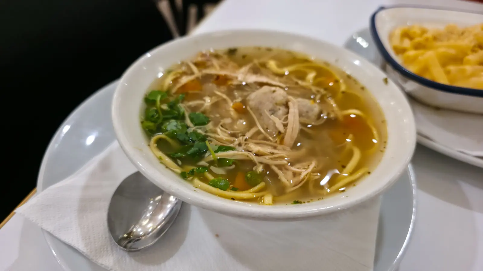 A bowl of chicken matzo ball and noodle soup garnished with herbs at the Brass Rail in Selfridges, a dish of macaroni cheese behind