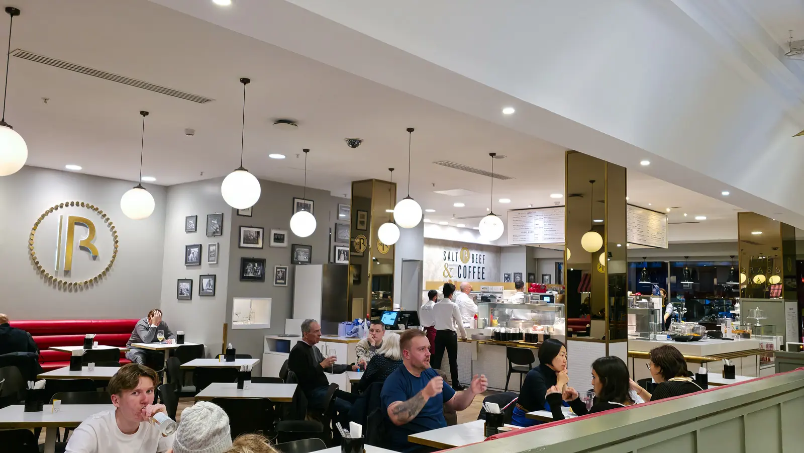 The dining room of the Brass Rail in Selfridges Food Hall, with diners at white tables, globe pendant lights and the gold BR logo on the wall