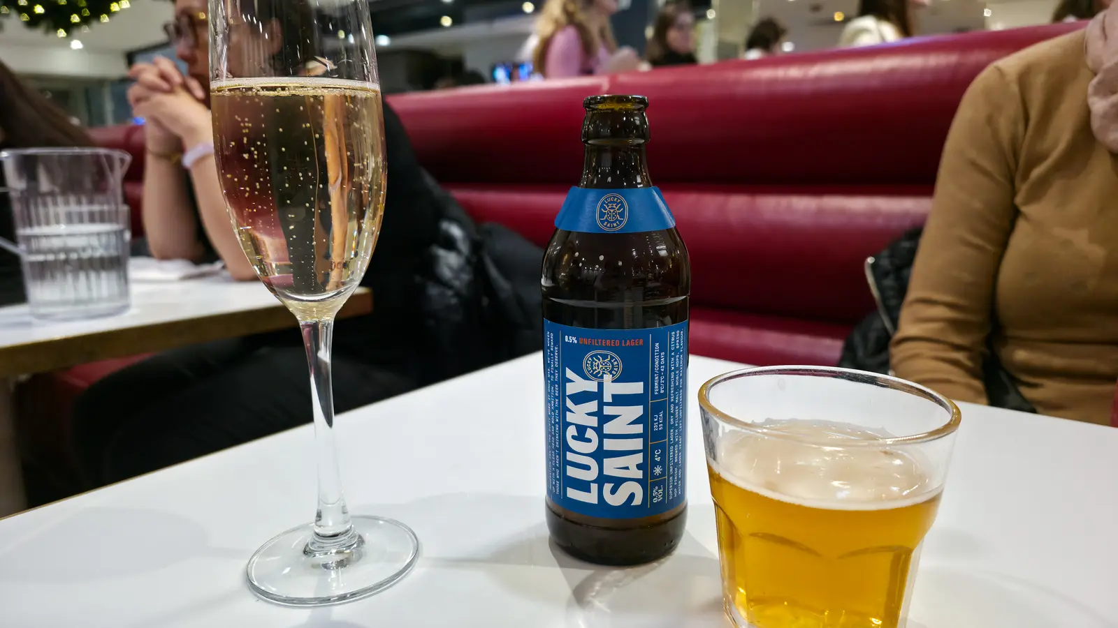 A bottle and glass of Lucky Saint alcohol-free lager beside a flute of champagne on a table at the Brass Rail in Selfridges