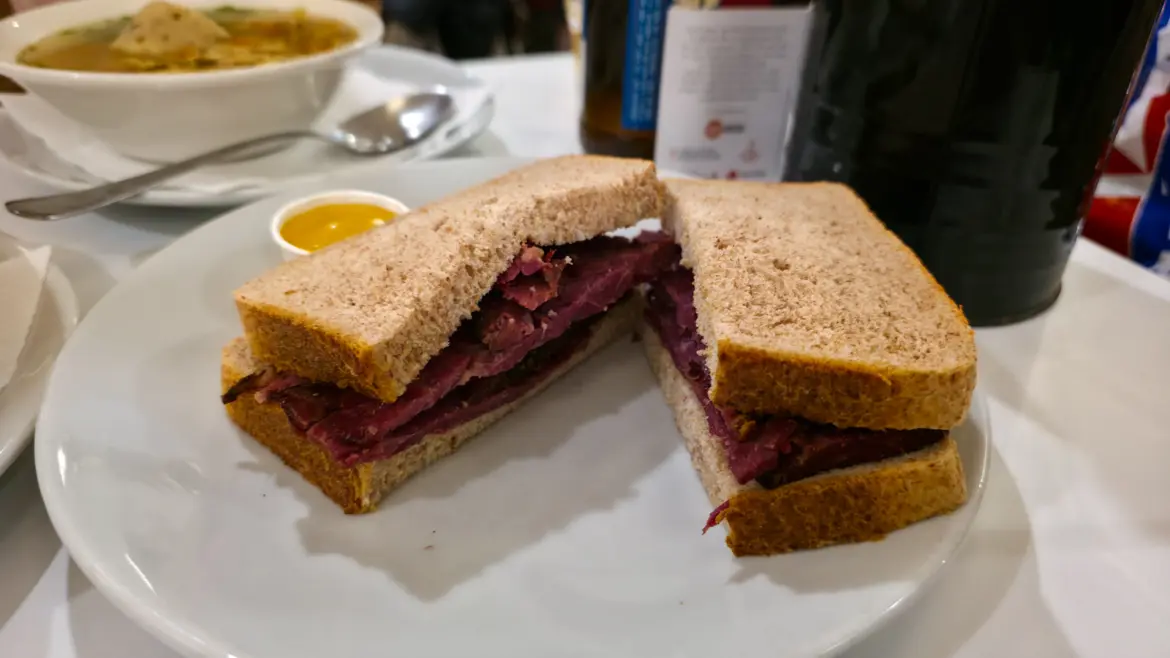 A pastrami sandwich on brown bread cut in half on a white plate with a pot of mustard, a bowl of chicken soup behind, at the Brass Rail in Selfridges