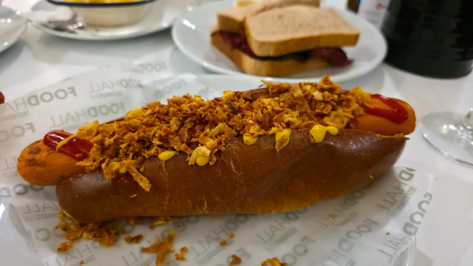 A vegan hot dog in a brioche bun with crispy onions, ketchup and mustard at the Brass Rail, a pastrami sandwich on brown bread behind