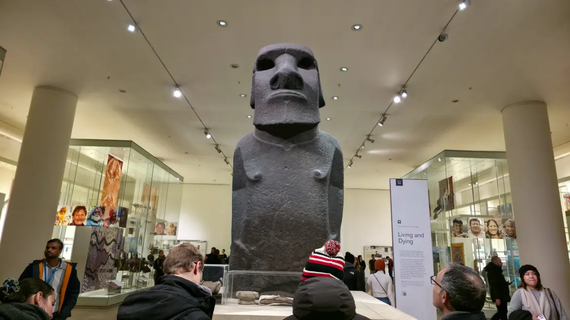 The basalt Moai Hoa Hakananai'a standing in the Living and Dying gallery of the British Museum, the carved figure with its iconic flat-topped head, deep-set eye sockets, prominent jaw, and hands curled at the front, with track lighting in a curving line around the white ceiling overhead and display cases of Pacific cultural material flanking it