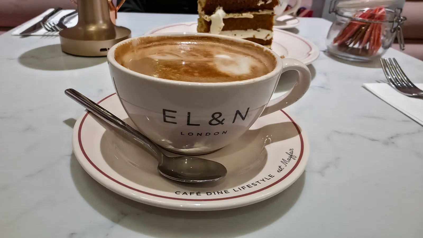 A flat white in an EL&N London cup and saucer on a marble table, cake in the background