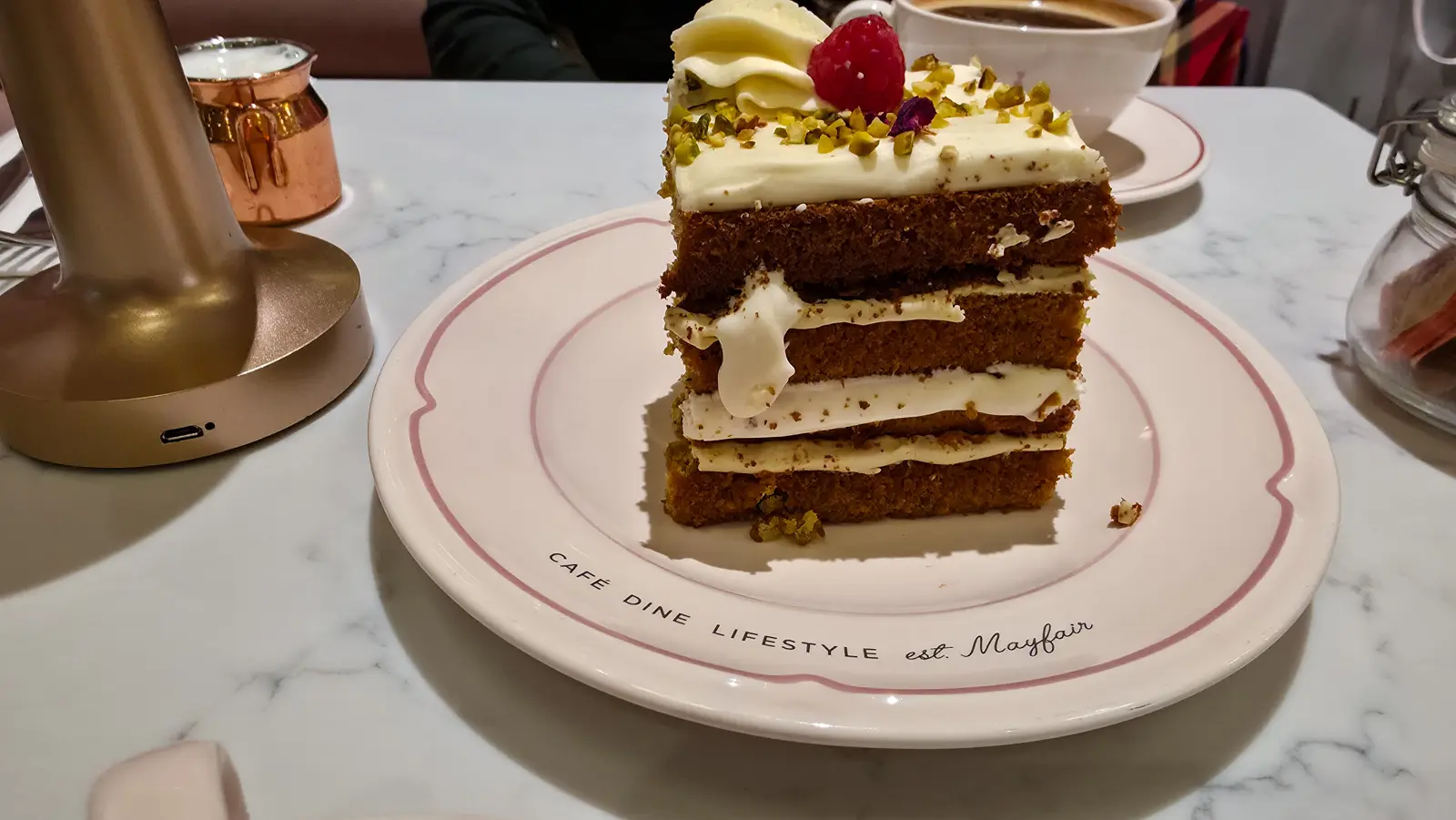 Tall slice of EL&N's gluten-free Pistachio & Rose Cake on an EL&N London plate, coffee behind