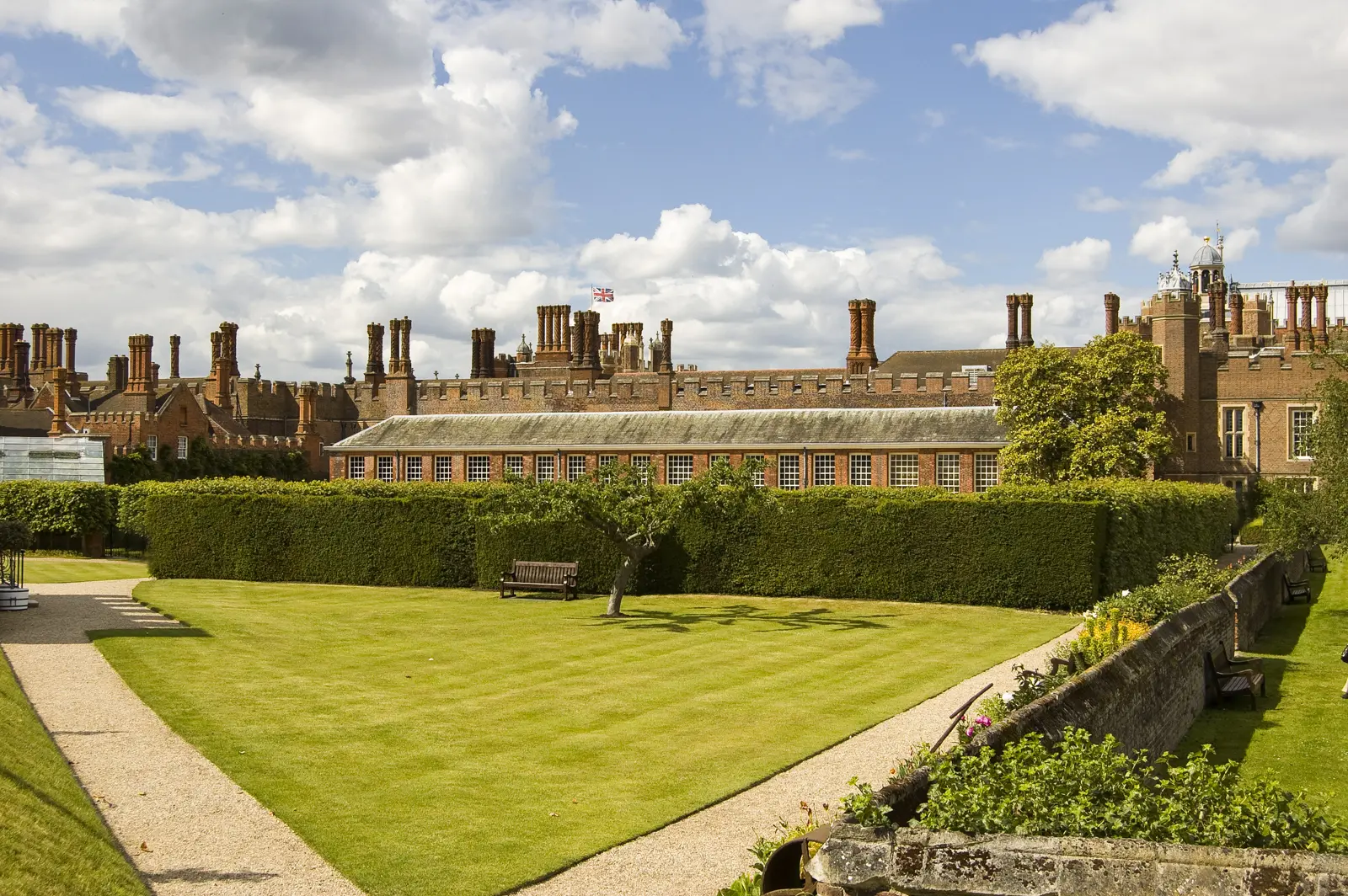 Hampton Court Palace from the gardens, its roofline crowded with ornate twisted Tudor chimneys above a clipped hedge and striped lawn