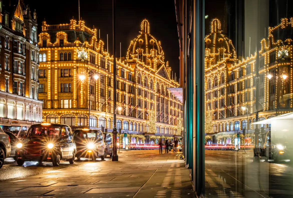 Harrods on Brompton Road at night with the entire terracotta façade outlined in golden lights, the central dome lit dramatically against the black sky, a Christmas tree illuminated at the entrance, black London taxis lining the street in the foreground, and the wet pavement reflecting the lights from a December evening shower