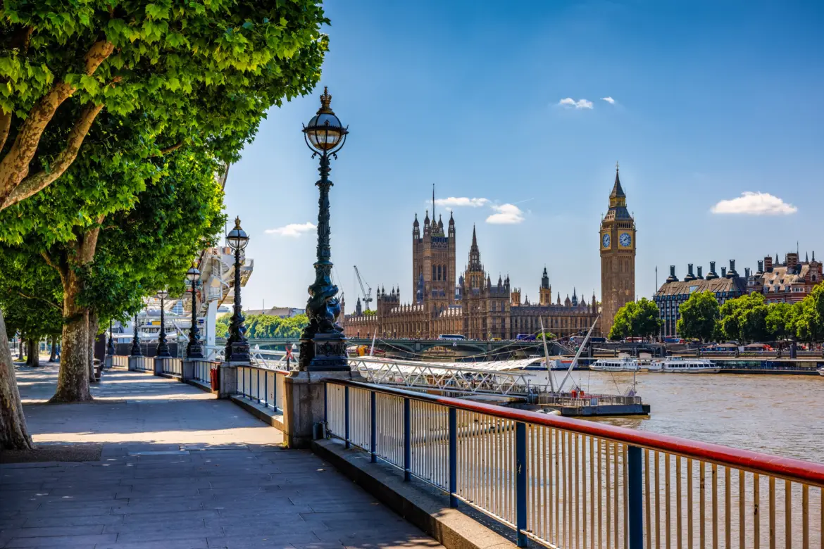 Tree-lined South Bank riverside walk with an ornate Victorian lamppost, looking across the Thames to the Houses of Parliament and Big Ben