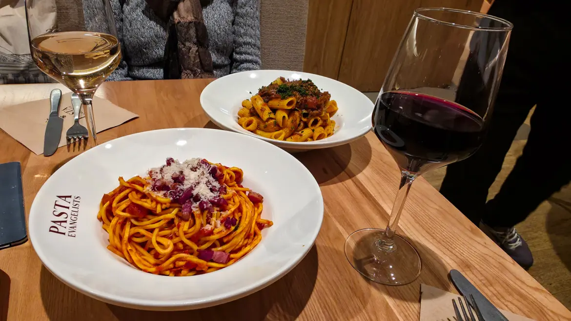 Two bowls of fresh pasta at Pasta Evangelists, New Oxford Street, with a glass of red and a glass of white wine on a wooden table.
