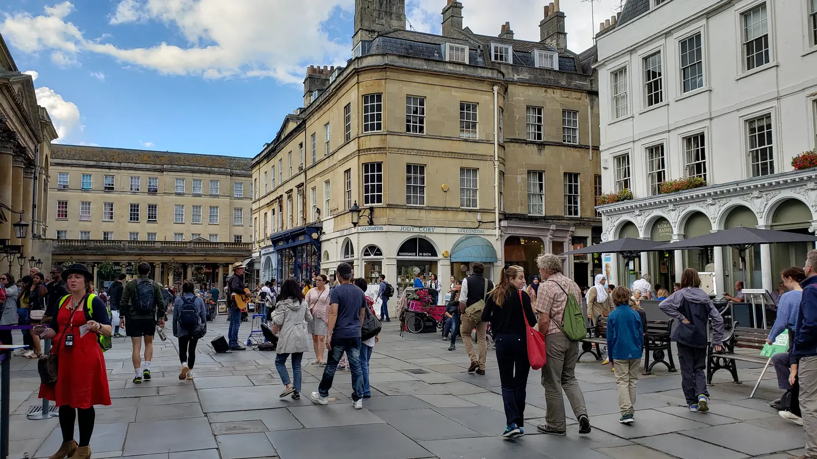 A busy paved square outside the Roman Baths in Bath, with shops, a busker and people among honey-coloured Georgian stone buildings under a blue sky