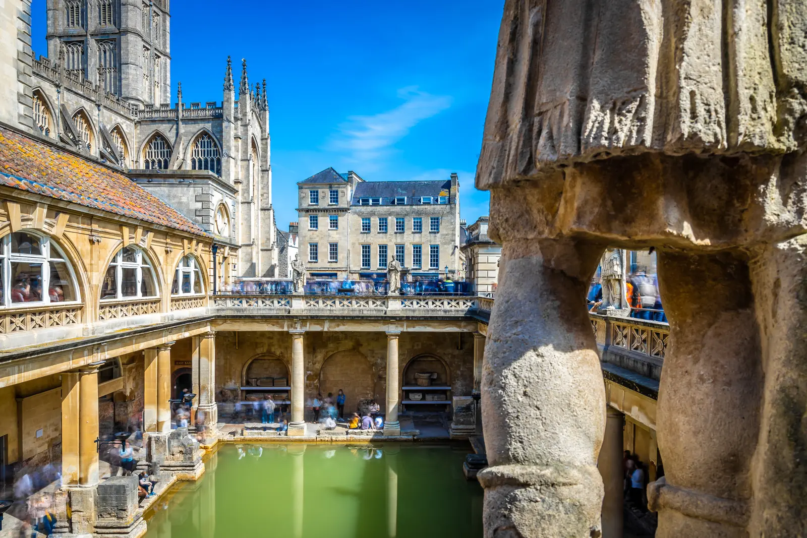 The green Great Bath at the Roman Baths in Bath with the pinnacled tower of Bath Abbey rising behind under a blue sky