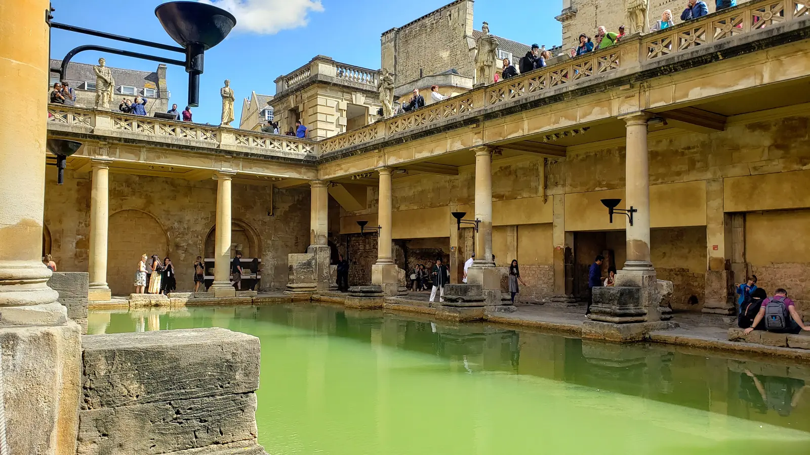 The Great Bath at the Roman Baths in Bath at water level, stone columns and a terrace of statues above against a blue sky