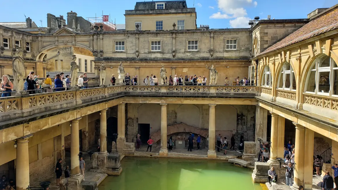 The Great Bath at the Roman Baths in Bath seen from the terrace, green water below a Victorian balustrade lined with statues, honey-coloured buildings and blue sky behind