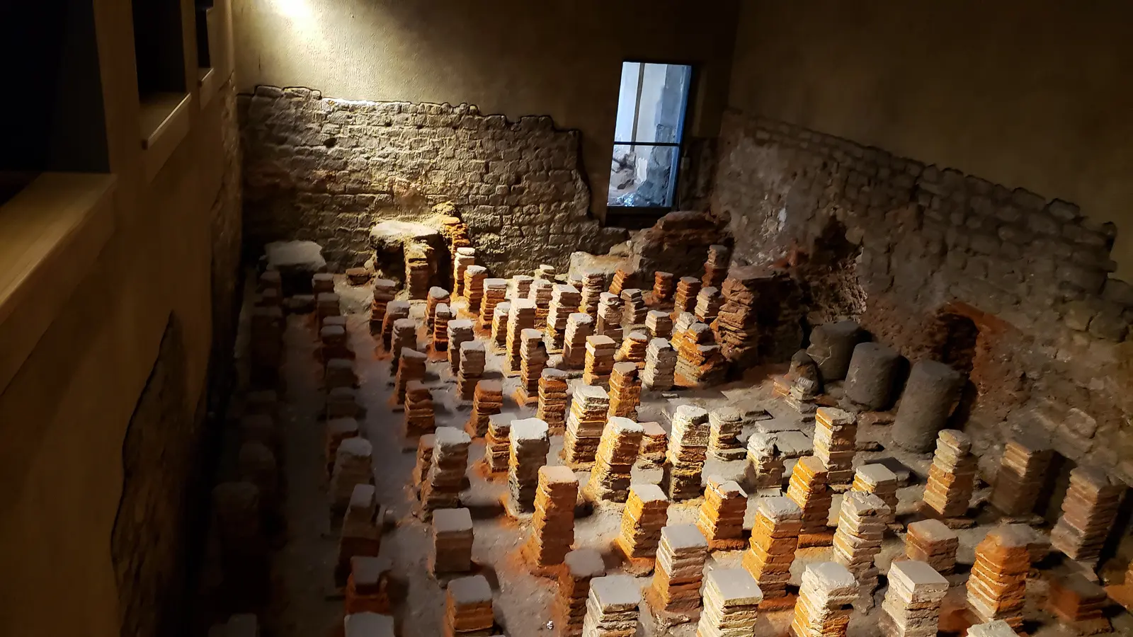 Rows of stacked brick and tile pillars under low warm light at the Roman Baths in Bath, the remains of a Roman hypocaust underfloor heating system