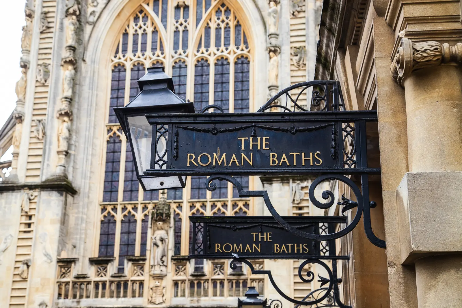A black wrought-iron sign reading The Roman Baths with a lantern, in front of the Gothic traceried window of Bath Abbey