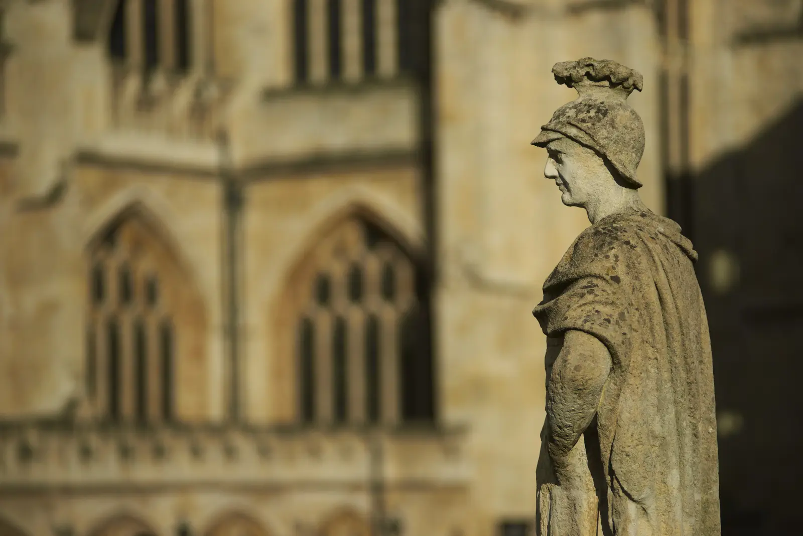 A weathered Victorian stone statue of a helmeted Roman figure in profile on the terrace at the Roman Baths, the Gothic stonework of Bath Abbey blurred behind