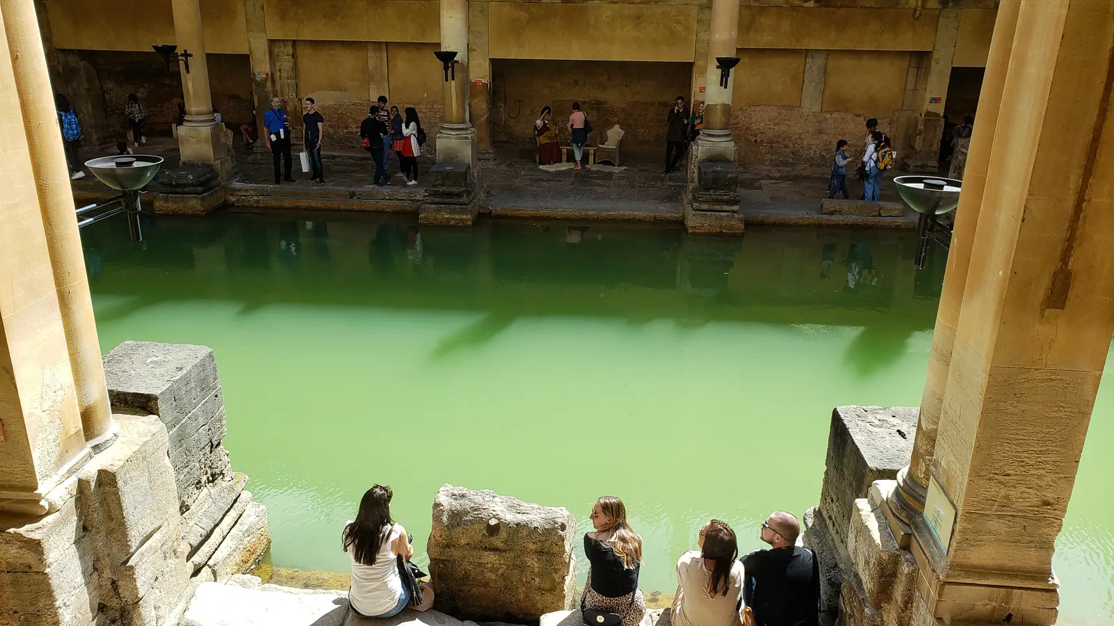 Visitors sitting on stone steps at the edge of the green Great Bath at the Roman Baths in Bath, columns and other visitors across the water