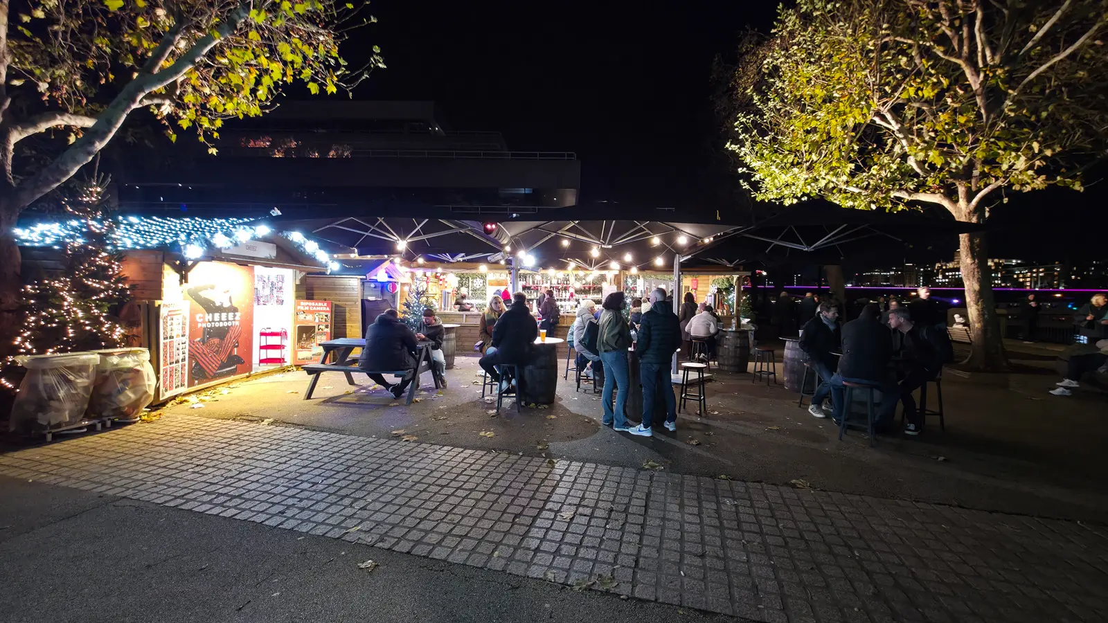 People standing at barrel tables under festoon lights beside wooden chalets at the Southbank Centre Winter Market at night, the concrete Southbank Centre behind