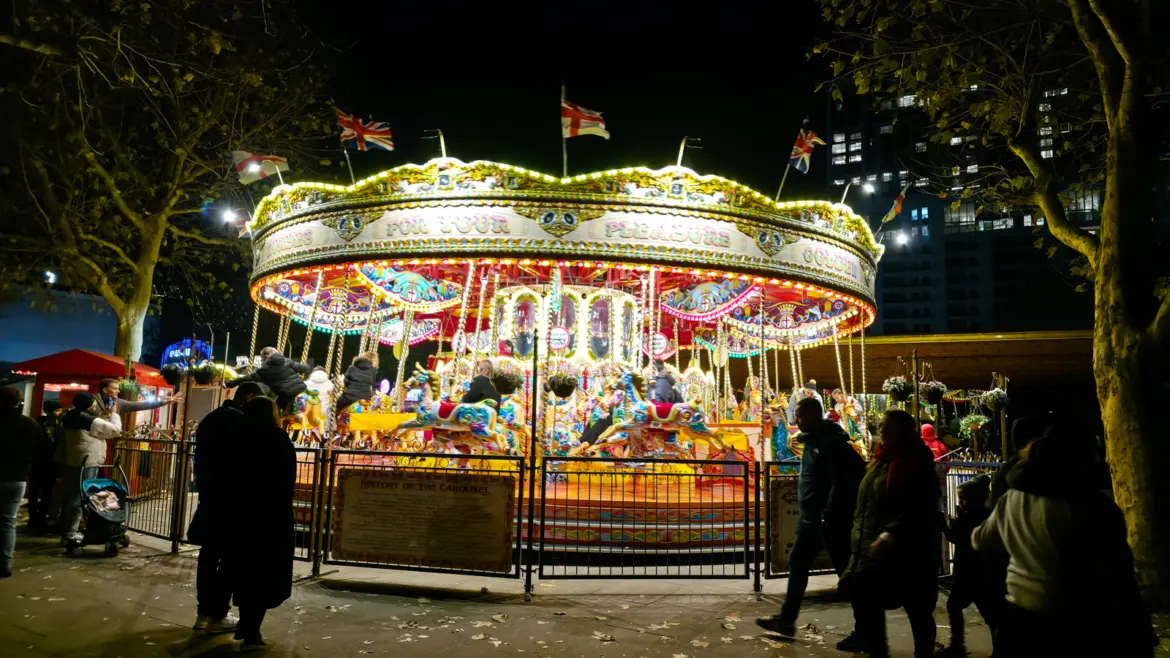An illuminated traditional carousel with painted horses and flags, lit gold at night, with crowds around it at the Southbank Centre Winter Market, London