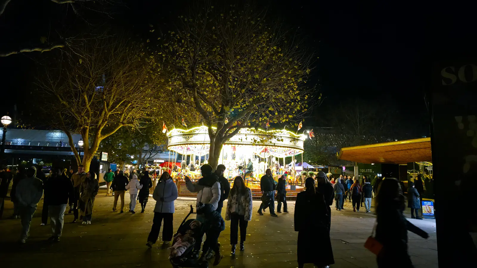 Crowds of visitors in winter coats at the Southbank Centre Winter Market at night, the gold-lit carousel glowing through bare trees behind them