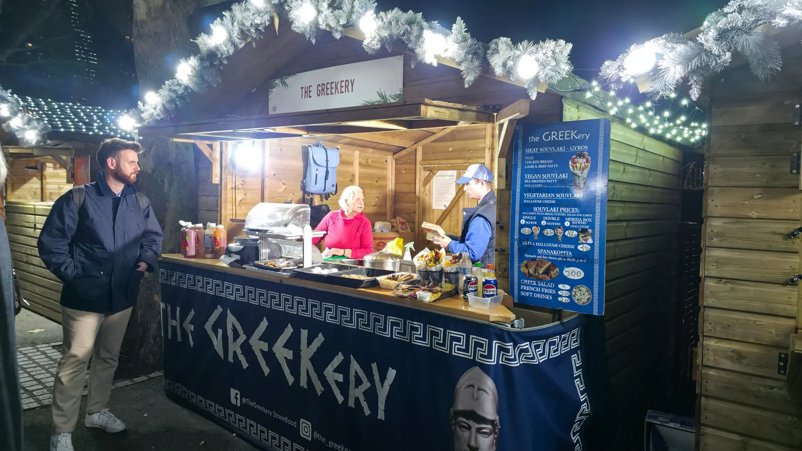 The Greekery, a wooden Greek street-food chalet lit up at night at the Southbank Centre Winter Market, its counter and menu board for souvlaki and gyros