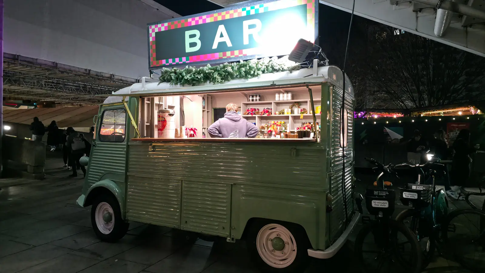 A sage-green vintage Citroen H van converted into a bar serving hot drinks, with an illuminated BAR sign, at the Southbank Centre Winter Market at night
