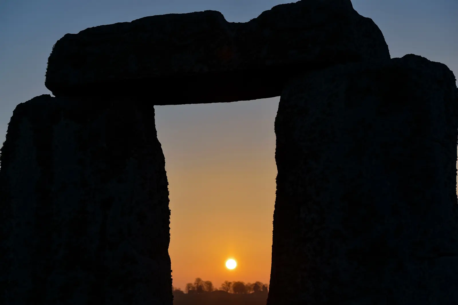 The sun setting through a Stonehenge trilithon, Wiltshire, the stones in black silhouette against an orange sky