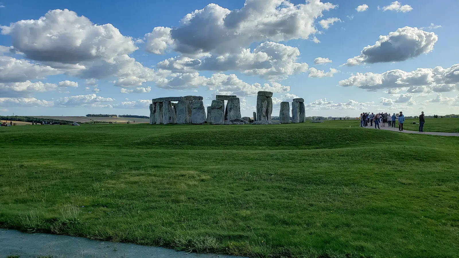 Stonehenge across a wide green field, Wiltshire, under a big blue sky, with visitors gathered on the path to one side