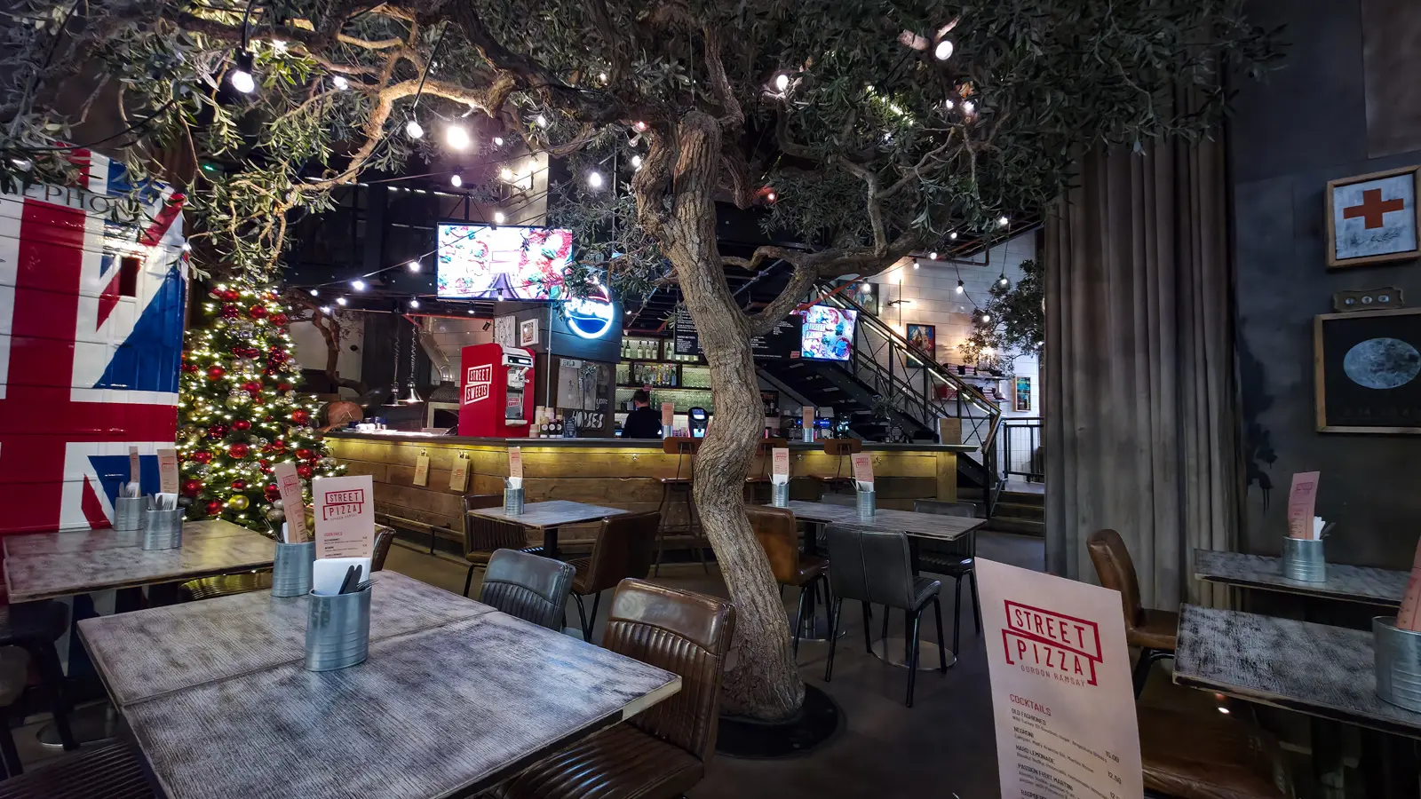 Inside Street Pizza by Gordon Ramsay, London, a full-size olive tree rises through the dining room under strings of festoon lights