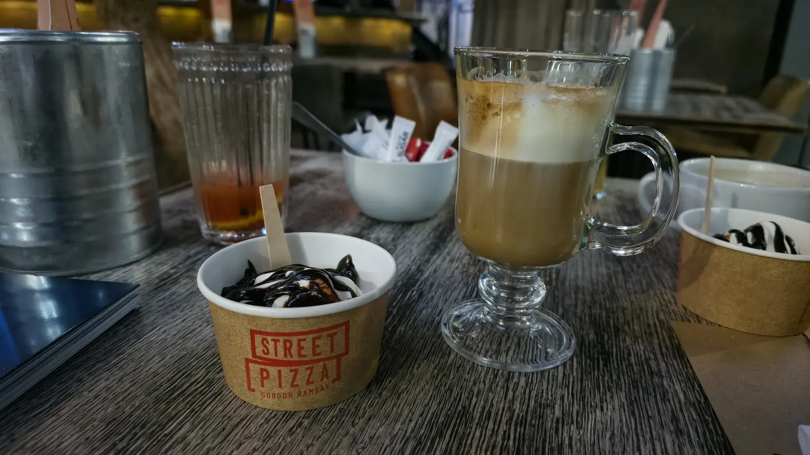 A cup of soft-serve with chocolate sauce at Street Pizza by Gordon Ramsay, London, beside a latte