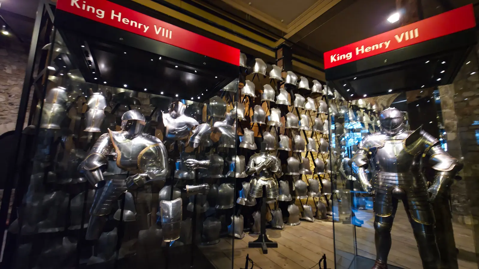 Suits of Henry VIII’s armour in glass cases beneath red King Henry VIII signage, with a wall of massed helmets behind, in the White Tower