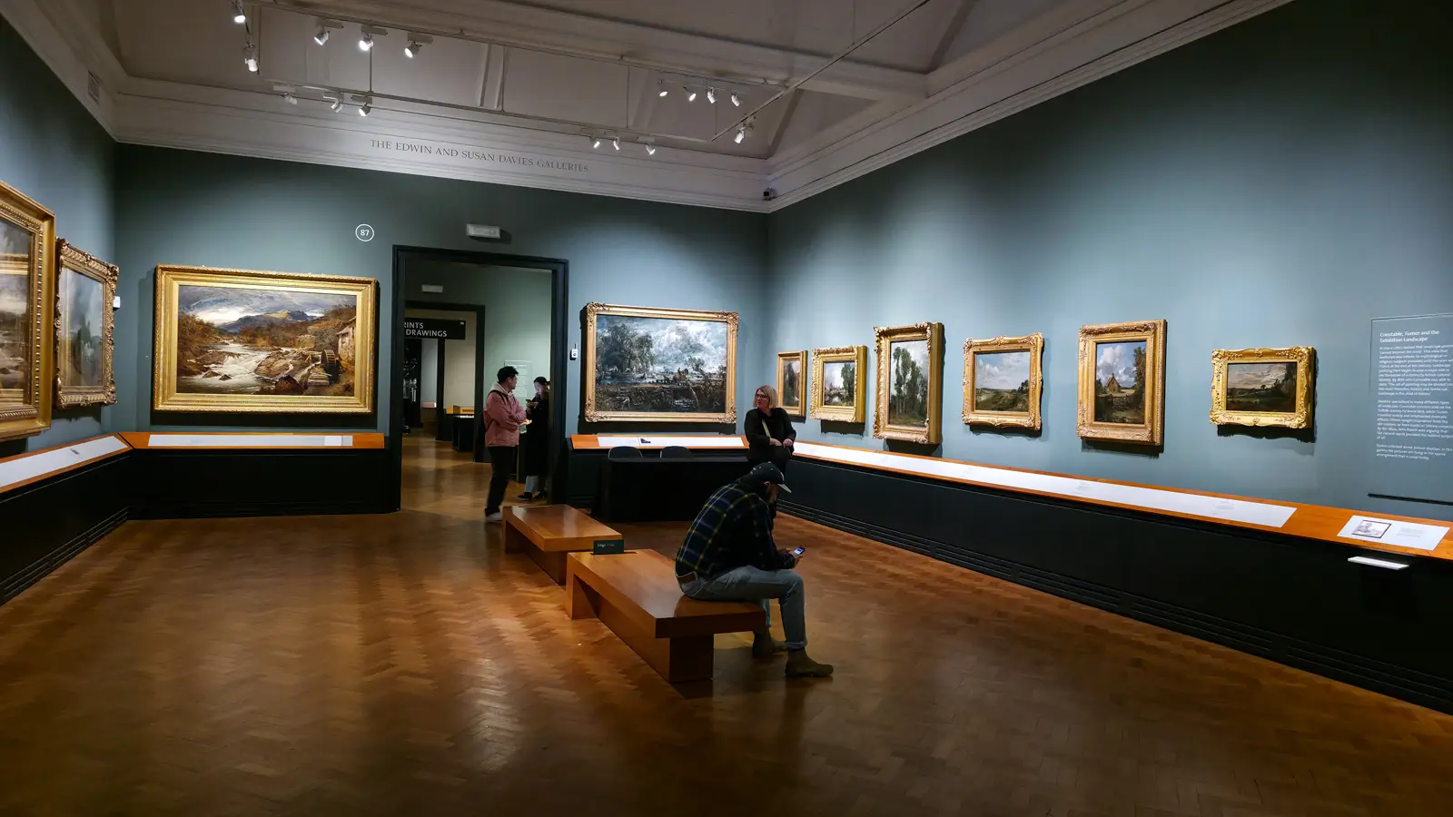 A gallery of British landscape paintings by Constable and Turner in gilt frames on teal walls, with visitors on benches, at the V&A, London