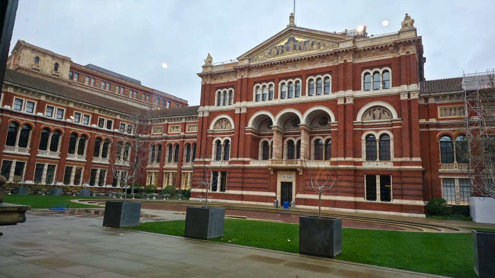 The red-brick and terracotta Victorian facade of the V&A around the John Madejski Garden courtyard, with a lawn and bare winter trees, on an overcast day