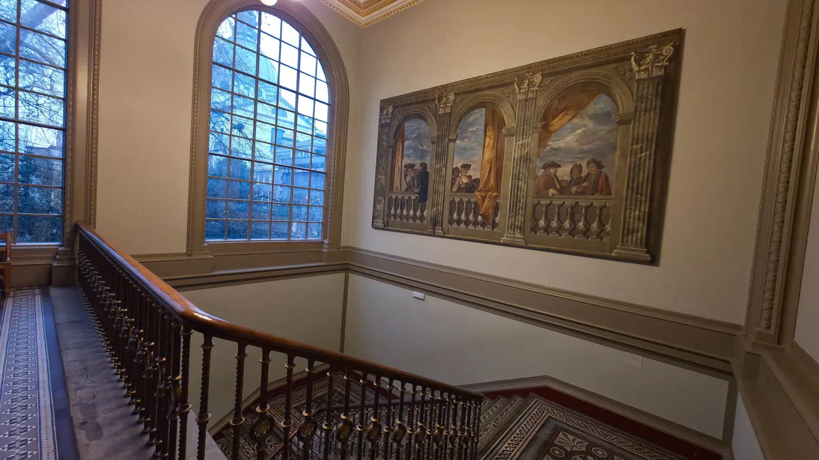 A painted and gilded staircase landing inside the V&A, with a large wall mural, a tall arched leaded window and a mosaic floor, London