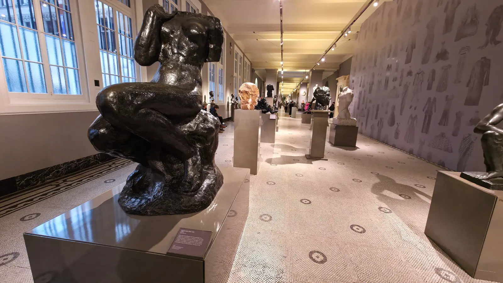 A dark bronze sculpture by Auguste Rodin on a reflective plinth in the V&A sculpture galleries, with more sculptures receding down the hall, London