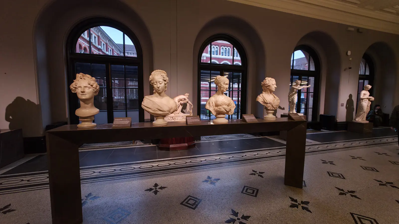 Rows of marble and terracotta portrait busts on a long plinth beside the arched windows of the V&A sculpture gallery, the courtyard garden visible behind, London