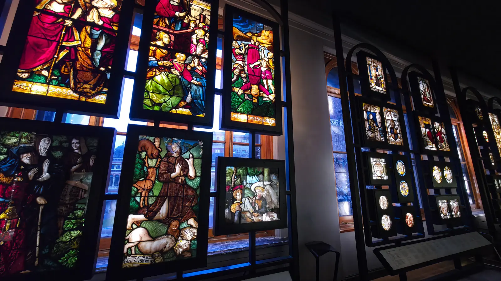 Backlit medieval and Renaissance stained glass panels glowing in black frames on a dark gallery wall at the V&A, London